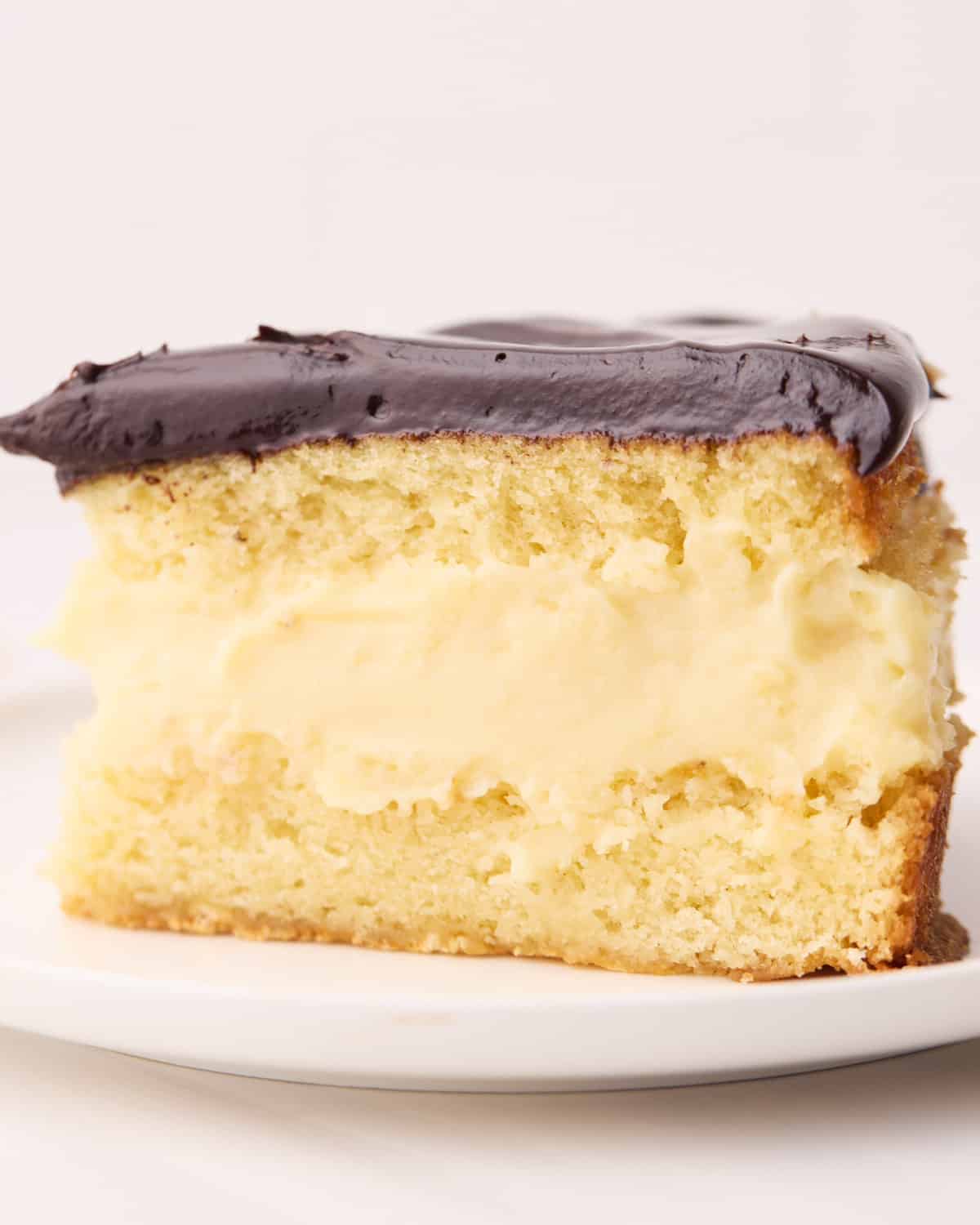 Close up of a slice of Boston cream pie showing the vanilla cake, pastry cream filling and chocolate ganache topping.