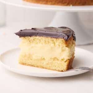 Slice of Boston cream pie on a plate with the full cake in the background - the distinct layers of sponge cake, pastry cream filling and chocolate glaze are visible.