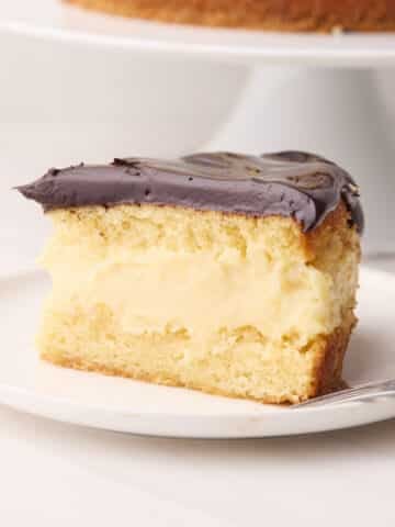 Slice of Boston cream pie on a plate with the full cake in the background - the distinct layers of sponge cake, pastry cream filling and chocolate glaze are visible.