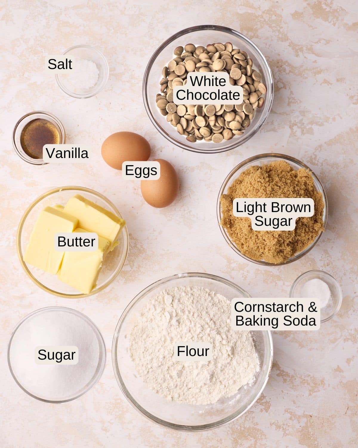 Labelled ingredients for white chocolate chip cookies, including salt, white chocolate chips, vanilla, eggs, light brown sugar, white sugar, butter, cornstarch, baking soda, and flour.