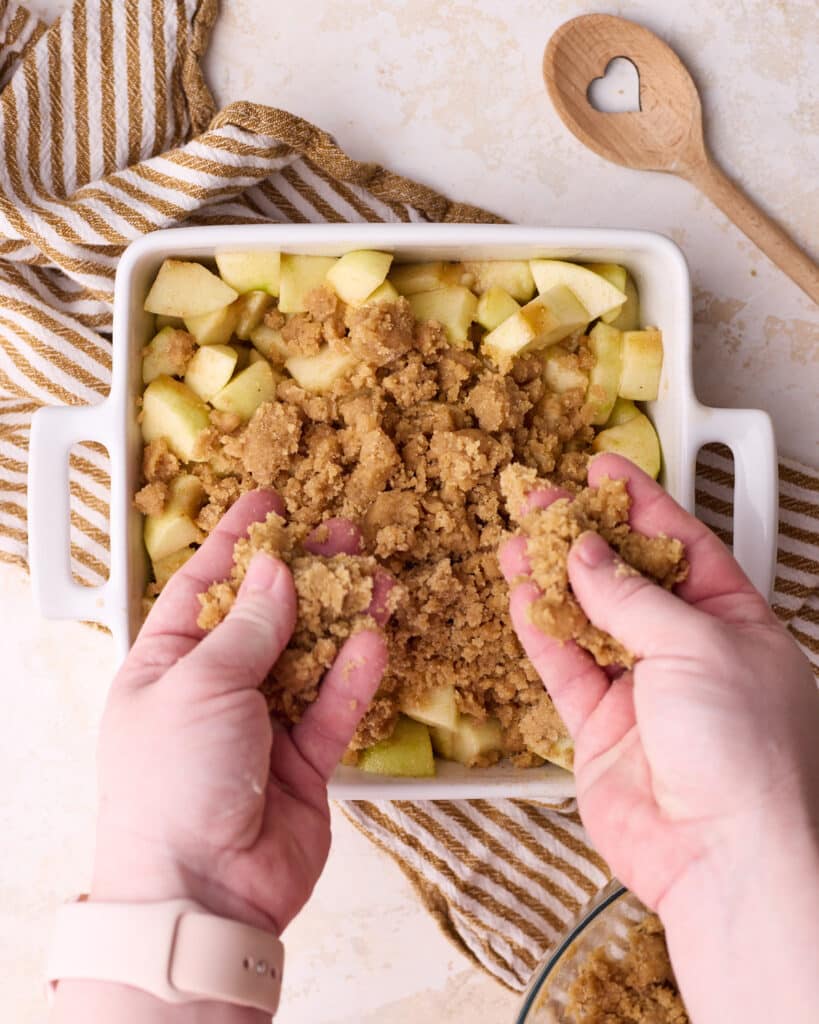 Apple crumble being assembled with apple filling on the bottom of a baking dish, and the crumb topping being sprinkled on top by hand.