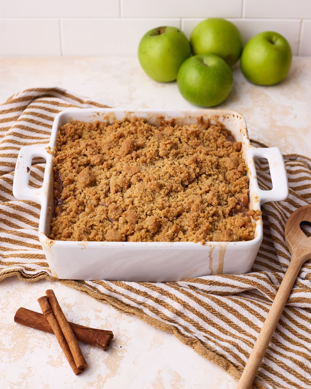 Freshly baked apple crumble in a baking dish with a golden crumb, surrounded by cinnamon sticks and fresh apples.