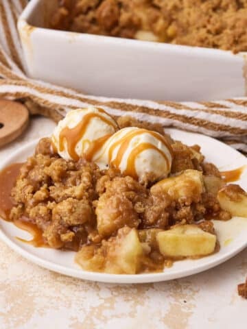 A portion of apple crumble on a plate dressed with 2 scoops of vanilla ice cream and a drizzle of caramel.