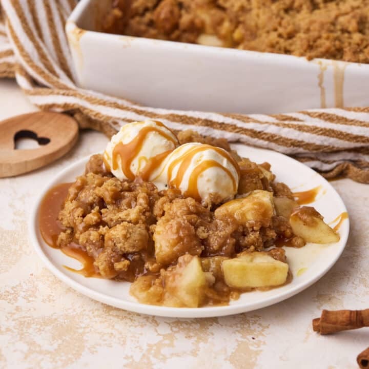 A portion of apple crumble on a plate dressed with 2 scoops of vanilla ice cream and a drizzle of caramel.