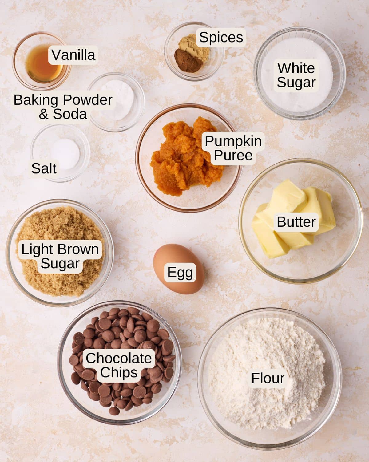 Ingredients for pumpkin chocolate chip cookies, including vanilla, leavening agents, spices, salt, white sugar, light brown sugar, pumpkin puree, butter, an egg, chocolate chips, and flour.