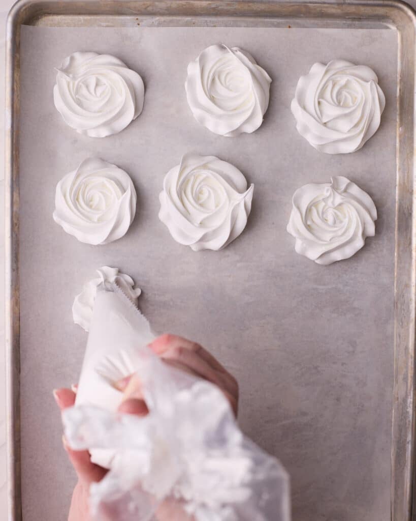 Meringue cookies being piped onto a prepared baking sheet in the shape of roses.