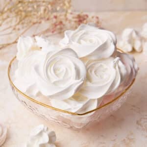 White rose meringue cookies in a love heart shaped glass bowl, showing off the beautiful designs.