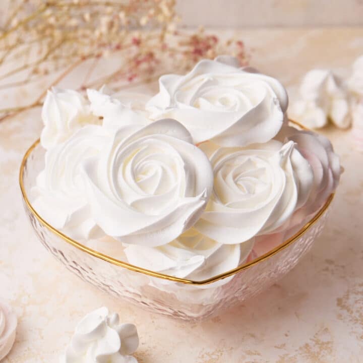White rose meringue cookies in a love heart shaped glass bowl, showing off the beautiful designs.