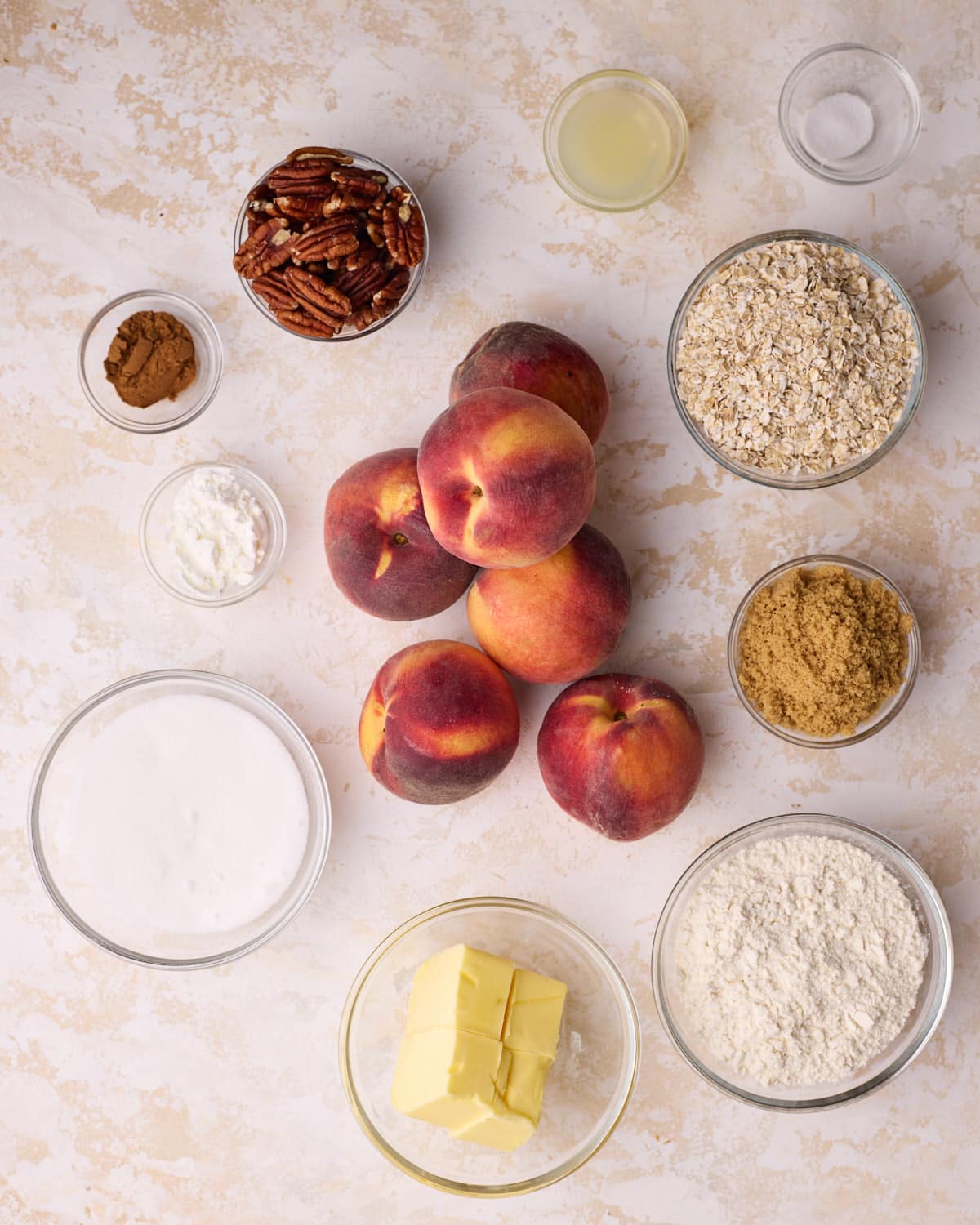 Ingredients to make peach crisp, including fresh peaches, butter, flour, white sugar, light brown sugar, oats, cinnamon, salt, lemon juice and pecans.