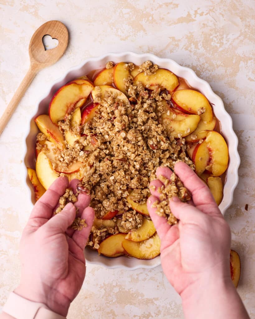 Crisp topping being sprinkled over a layer of fresh peaches in a round pie dish to make a peach crisp.