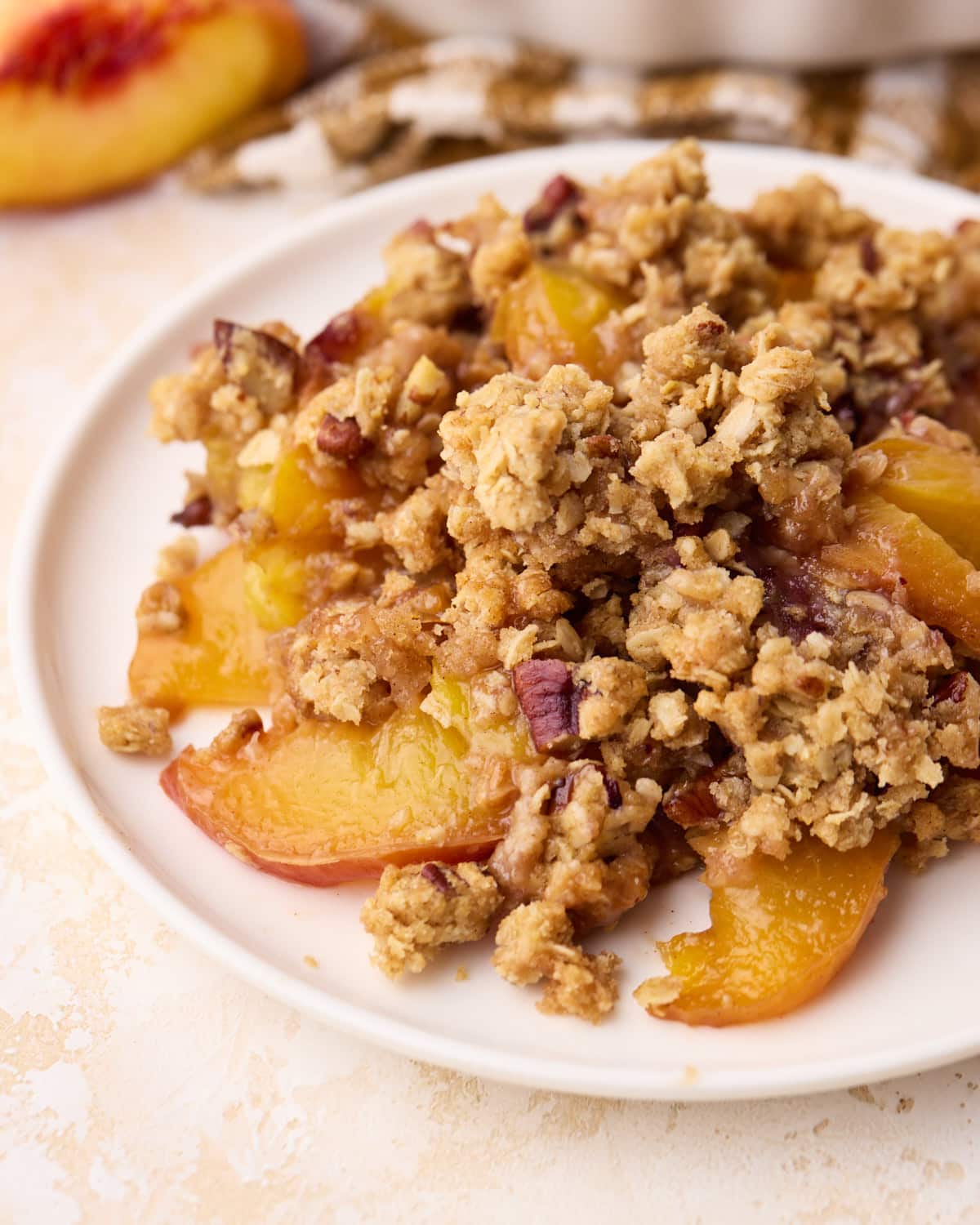 Close up of a serving of peach crisp on a plate with yellow peaches and red skins, under a crumbly topping with oats and pecans.