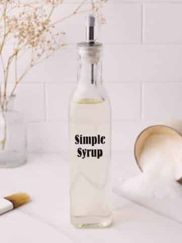 Simple syrup in a labelled glass bottle with a spout, surrounded by a fallen over cup of sugar and a pastry brush.