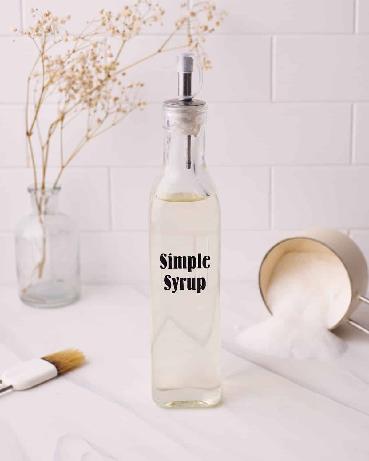 Simple syrup in a labelled glass bottle with a spout, surrounded by a fallen over cup of sugar and a pastry brush.