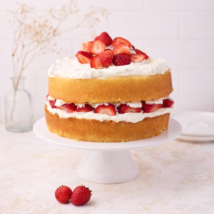 Vanilla sponge cake filled with strawberries and cream, on a white cake stand.