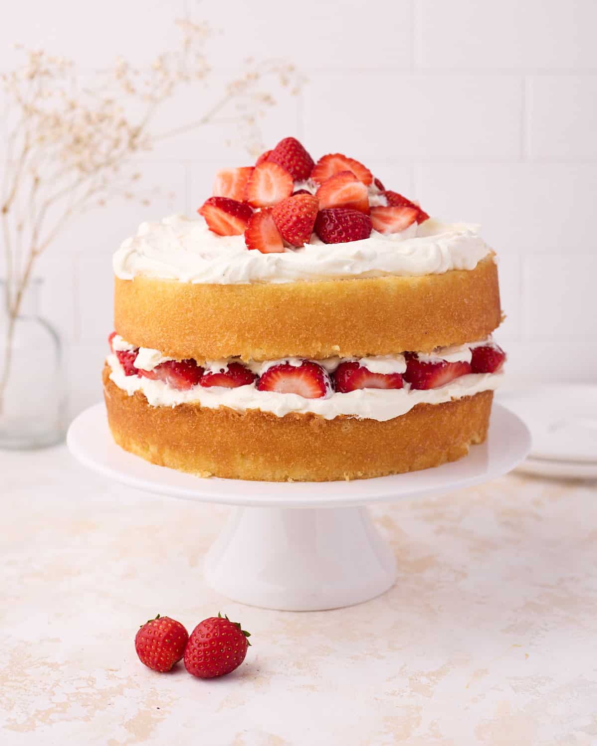Vanilla sponge cake filled with strawberries and cream, on a white cake stand. 