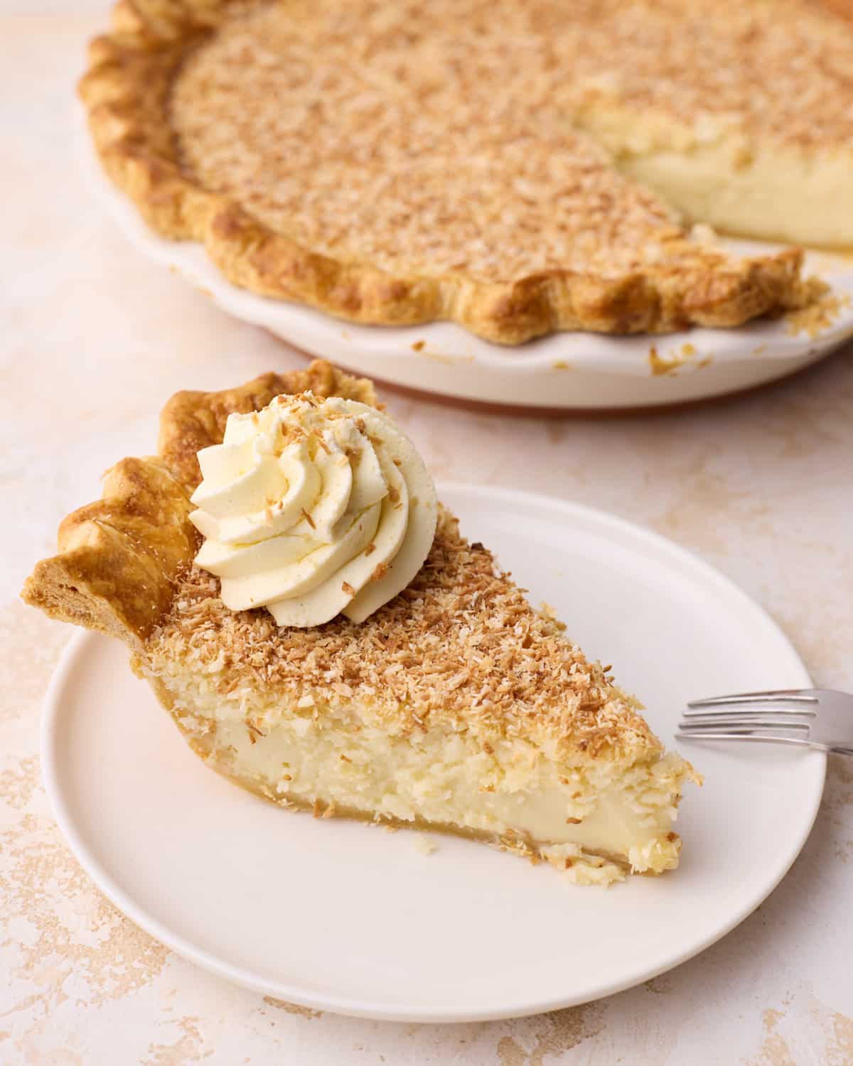 Overhead view of Side view of a slice of coconut custard pie on a plate with the rest of the pie in the background.