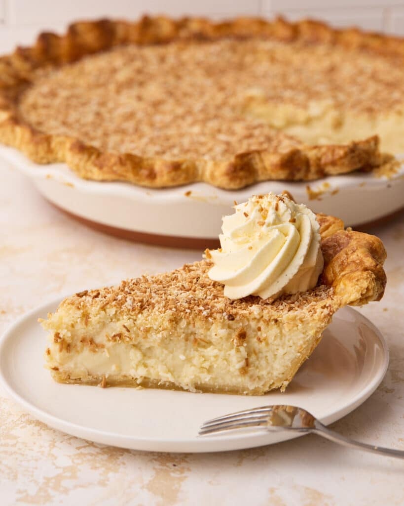 Side view of a slice of coconut custard pie on a plate with the rest of the pie in the background.