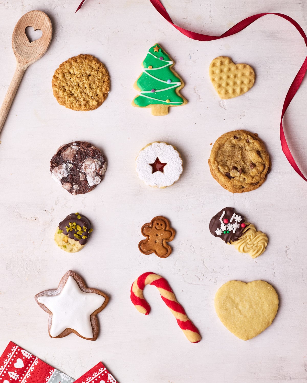 Each of the Chirstmas cookies spaced out evenly in 3 lines, with a wooden spoon, ribbon, and napkins around them for decoration.