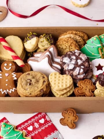 An open cookie box displaying all of the different christmas cookies, with festive decorations and spare cookies dotted around the outside.