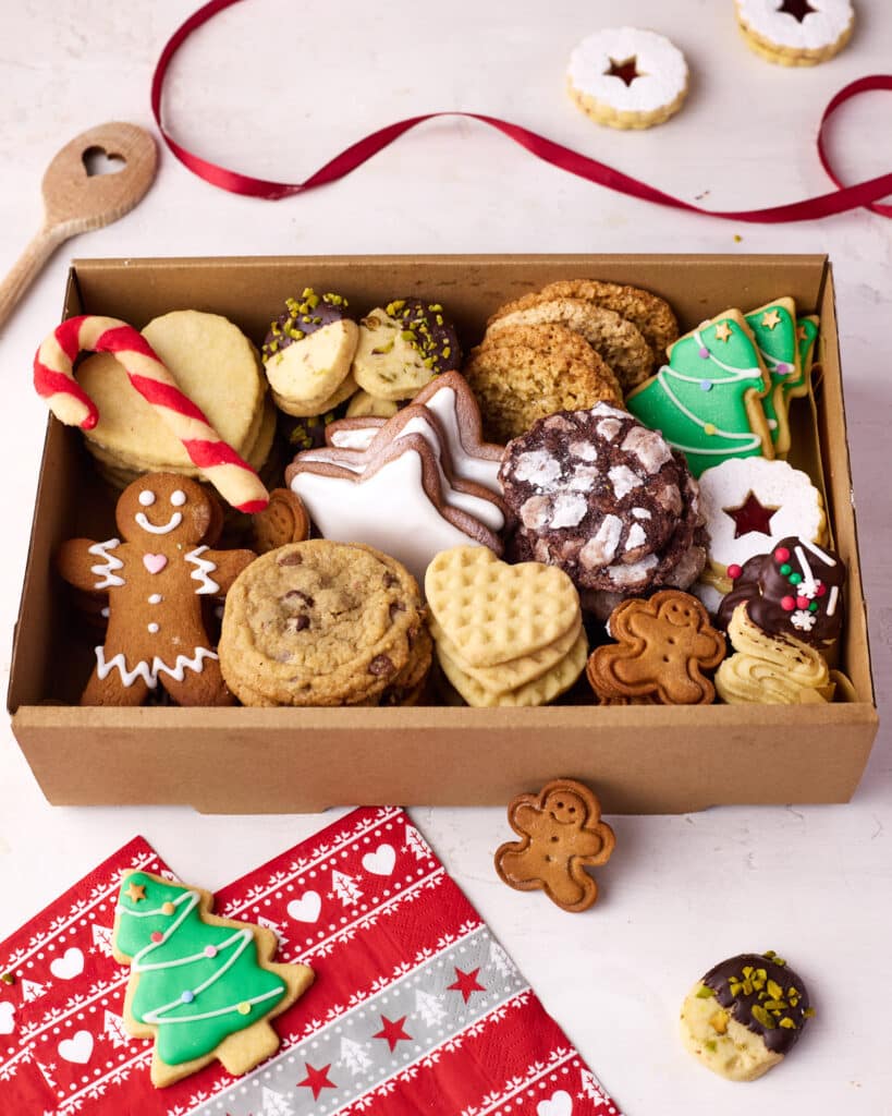 An open cookie box displaying all of the different christmas cookies, with festive decorations and spare cookies dotted around the outside.