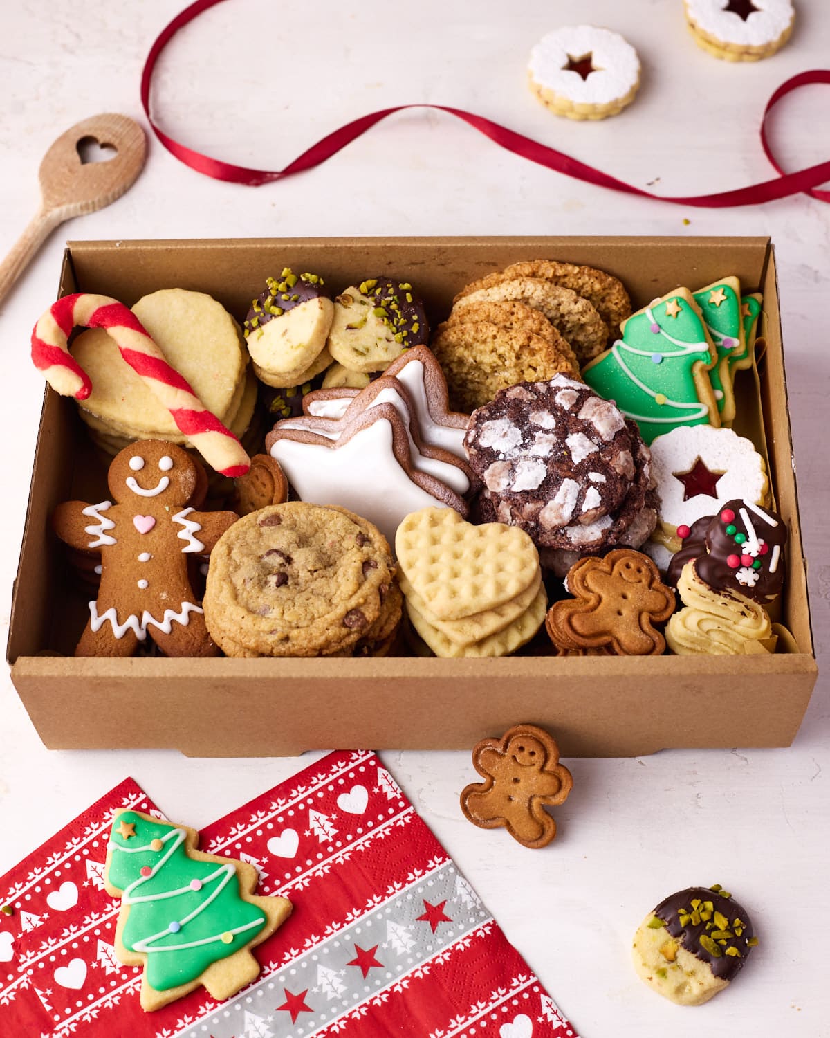 An open cookie box displaying all of the different christmas cookies, with festive decorations and spare cookies dotted around the outside.