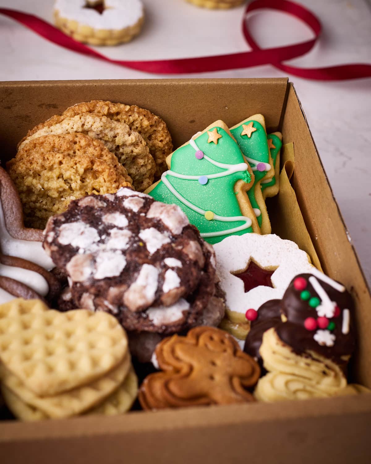A close up of inside the cookie box, displaying oatmeal lace cookies, Linzer cookies, Danish butter cookies, chocolate crinkle cookies, mini gingerbread men, sugar cookie Christmas trees, and love heart shortbread cookies.