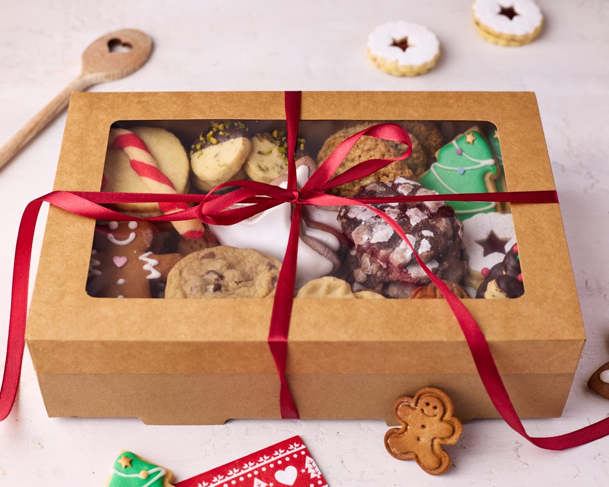 Christmas cookie box wrapped in red ribbon, with a wooden spoon and a few cookies dotted around it.