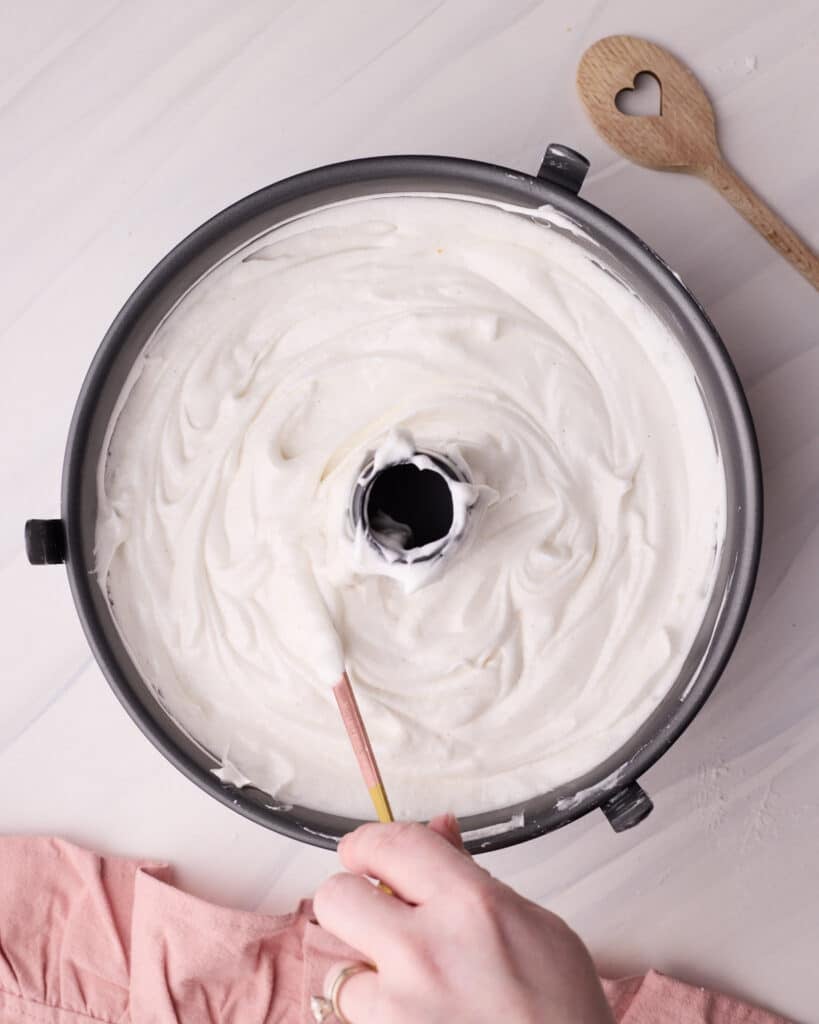 Angel food cake in a tube pan with a skewer being run through it to release air bubbles.