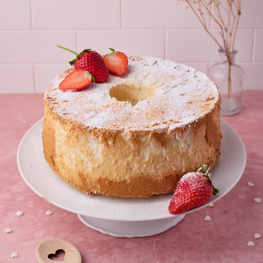 Angel food cake on a white cake stand with a dusting of icing sugar and fresh strawberries.