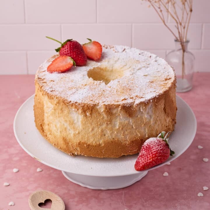 Angel food cake on a white cake stand with a dusting of icing sugar and fresh strawberries.