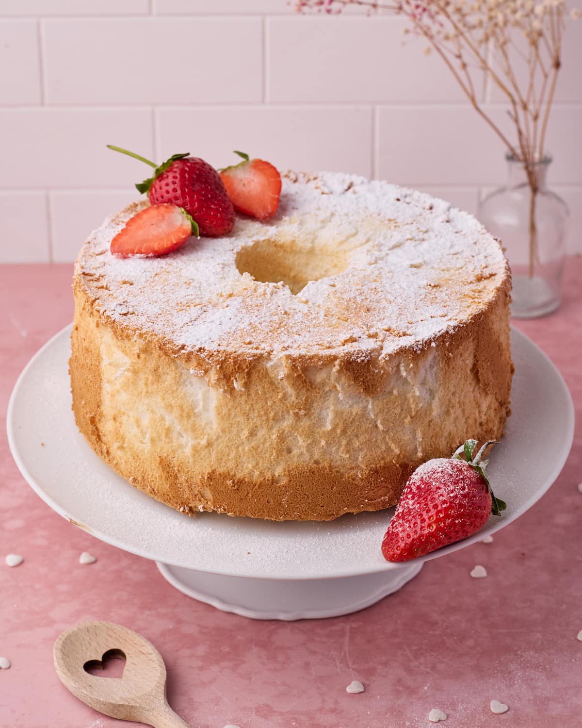Angel food cake on a white cake stand with a dusting of icing sugar and fresh strawberries. 