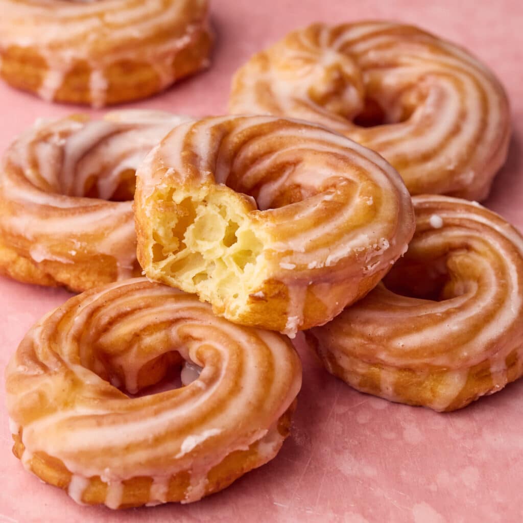 A pile of French crullers on a pink backdrop, one has a bite taken out of it to show the soft, honeycomb texture inside.