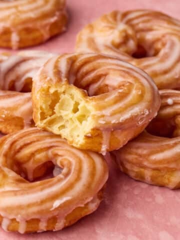 A pile of French crullers on a pink backdrop, one has a bite taken out of it to show the soft, honeycomb texture inside.