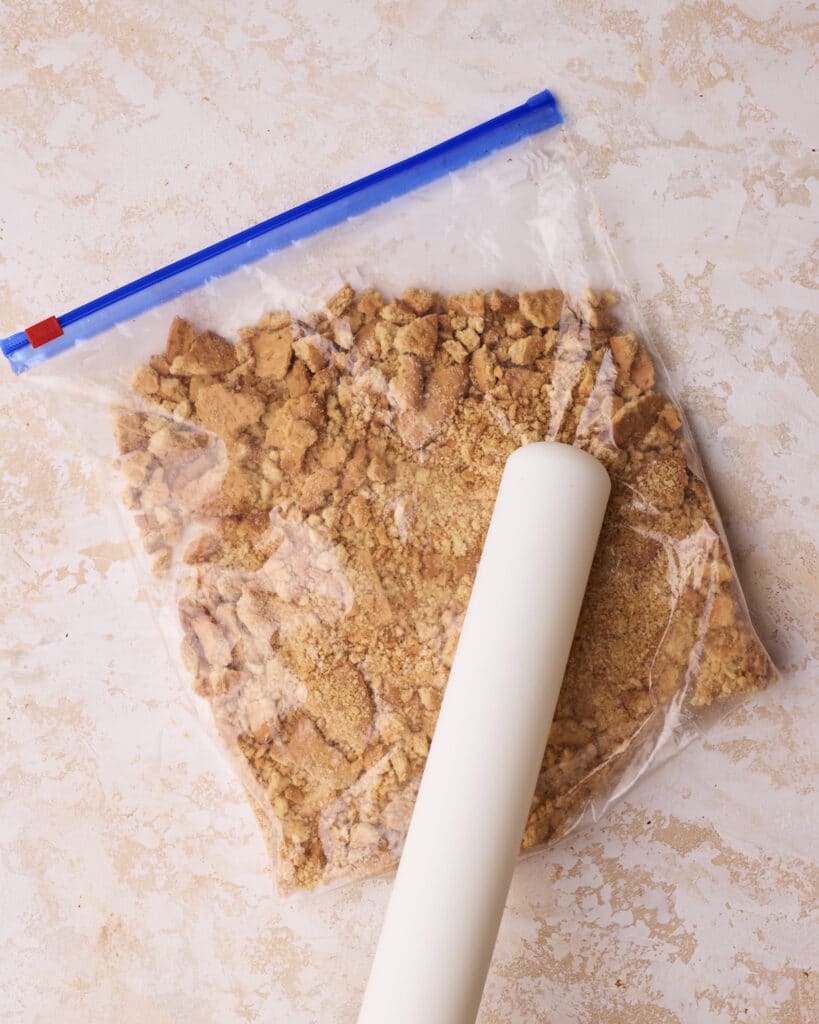 Digestive biscuits being crushed with a rolling pin in a plastic bag to make tiffin.