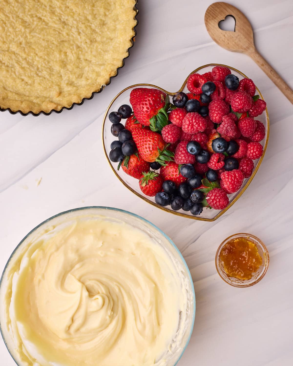 Ingredients to make fresh fruit tart including a tart shell, a bowl of fresh berries, including strawberries, raspberries and blueberries, some homemade pastry cream, and a small amount of apricot jam to glaze.