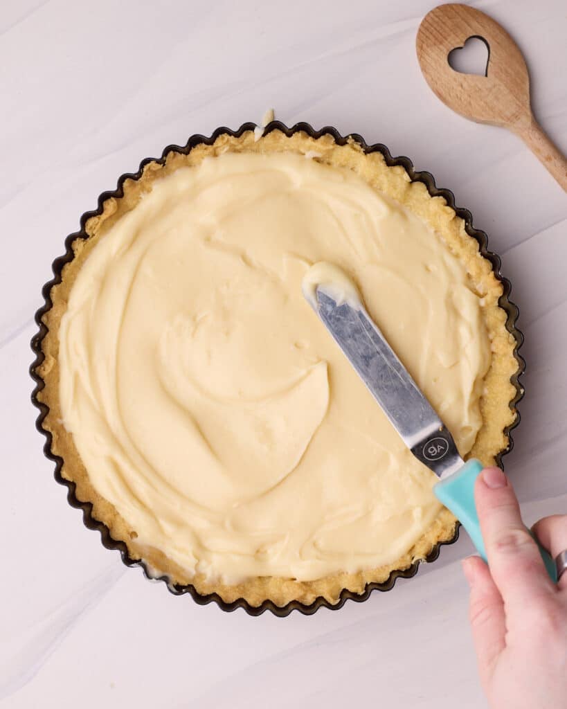 Spreading creamy pastry cream into a tart shell to make a fresh fruit tart.