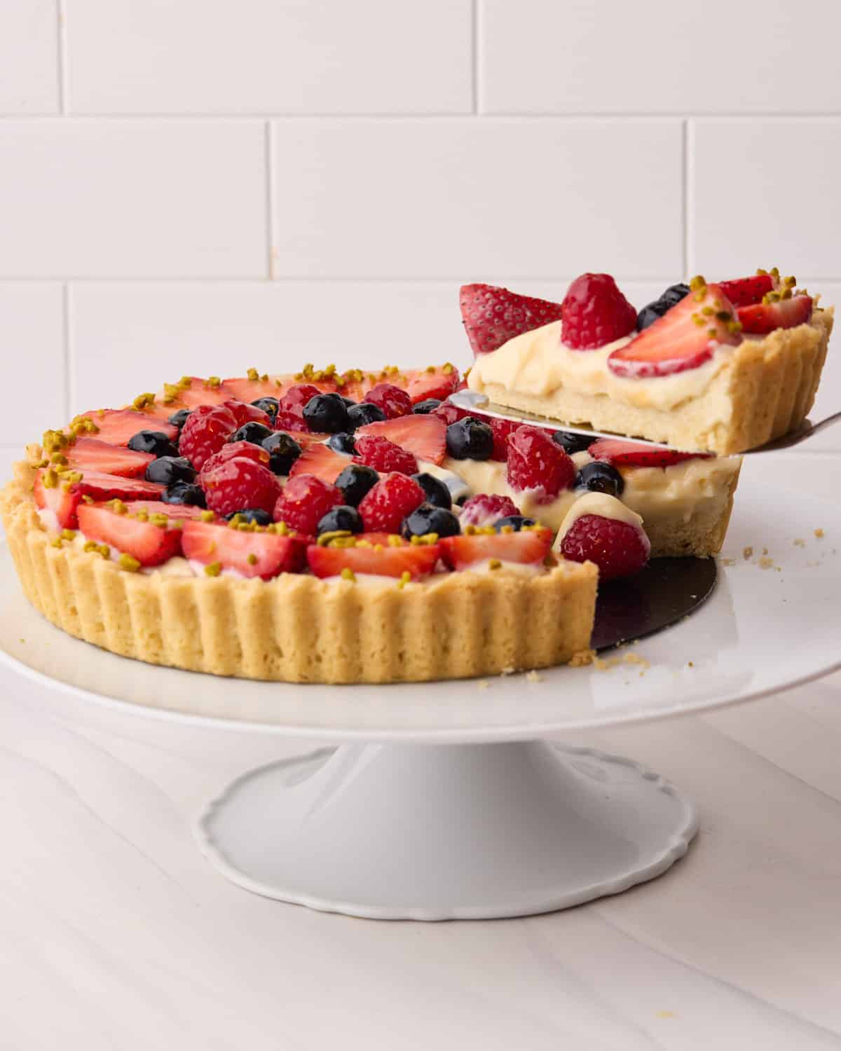 Fresh fruit tart with strawberries, raspberries, and blueberries, on a cake stand with a slice being taken out of it to show the creamy pastry cream filling.