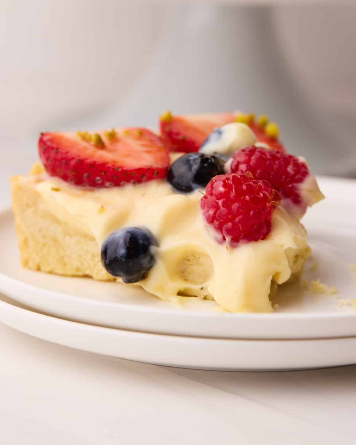Slice of fruit tart on a plate with creamy custard filling oozing over the side and fresh berries falling from the top.