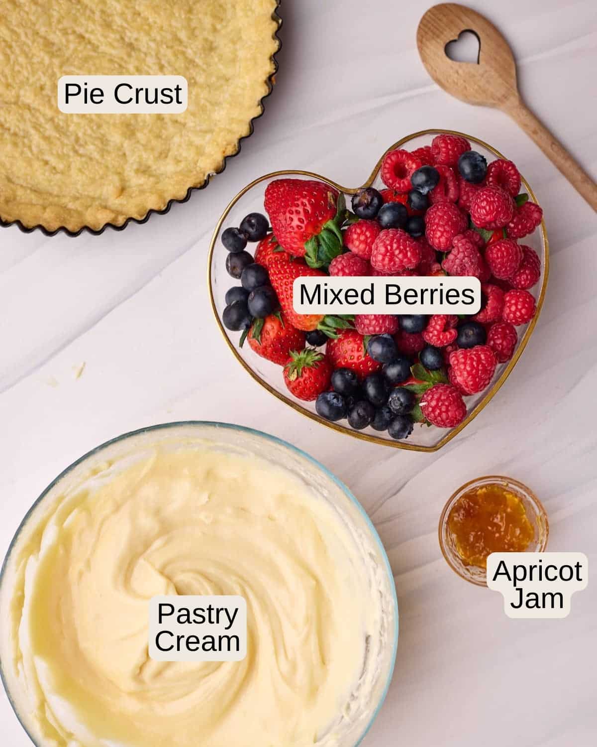 Ingredients to make fresh fruit tart including a tart shell, a bowl of fresh berries, including strawberries, raspberries and blueberries, some homemade pastry cream, and a small amount of apricot jam to glaze.