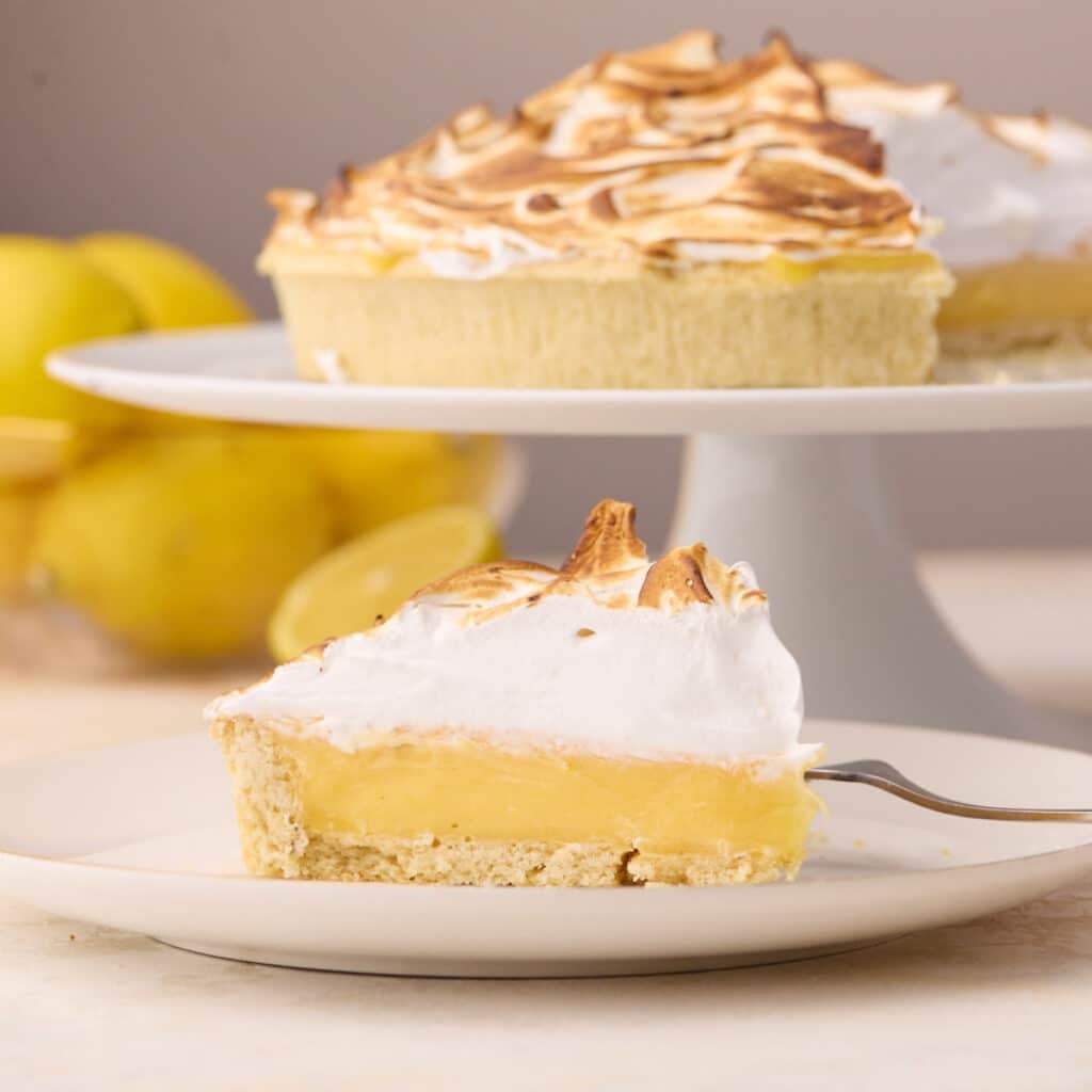Slice of lemon meringue pie on a plate with the whole pie in the background on a cake stand.