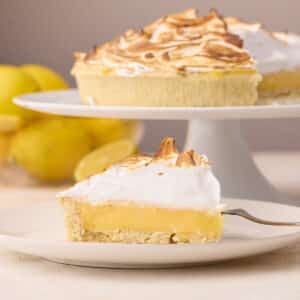 Slice of lemon meringue pie on a plate with the whole pie in the background on a cake stand.