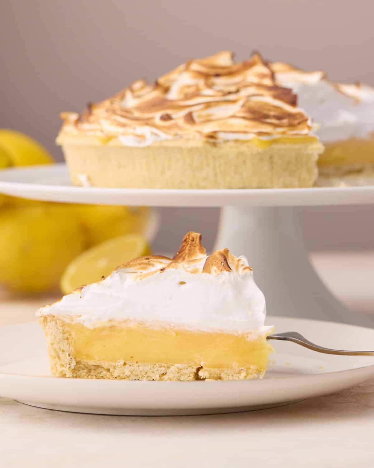 Slice of lemon meringue pie on a plate with the whole pie in the background on a cake stand.
