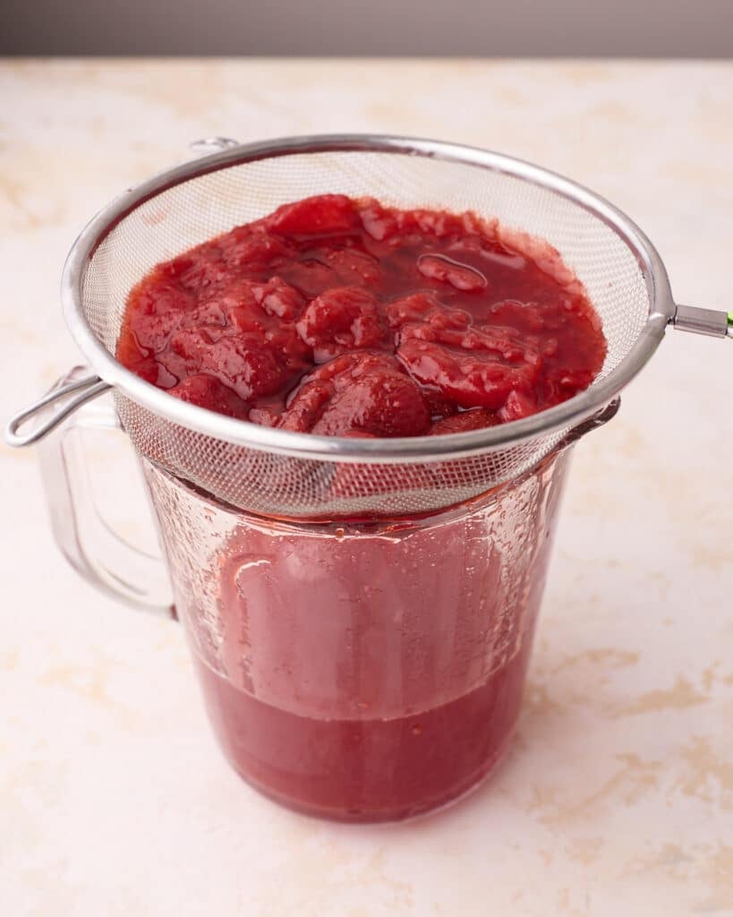 Strawberry simple syrup in a jug, being strained through a fine-mesh strainer to remove the strawberry solids.