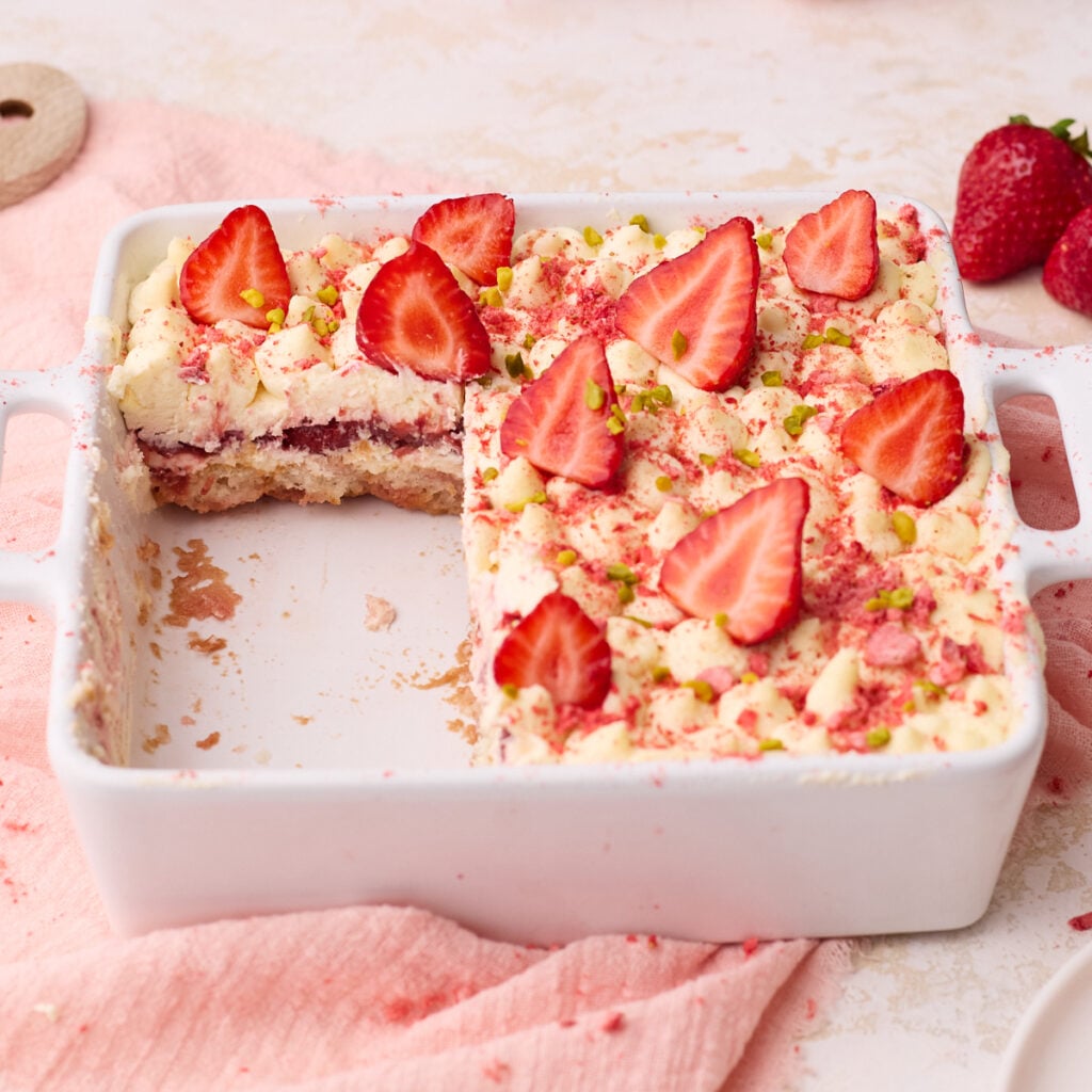 Strawberry tiramisu in a white baking dish with a few slices removed so you can see the layers inside of ladyfingers soaked in strawberry syrup, mascarpone cream and strawberry filling.