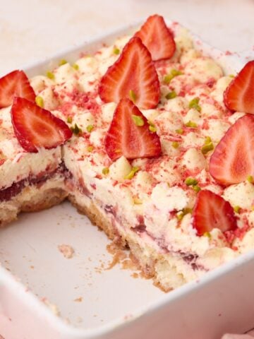 Strawberry tiramisu in a white baking dish with a few slices removed so you can see the layers inside of ladyfingers soaked in strawberry syrup, mascarpone cream and strawberry filling.