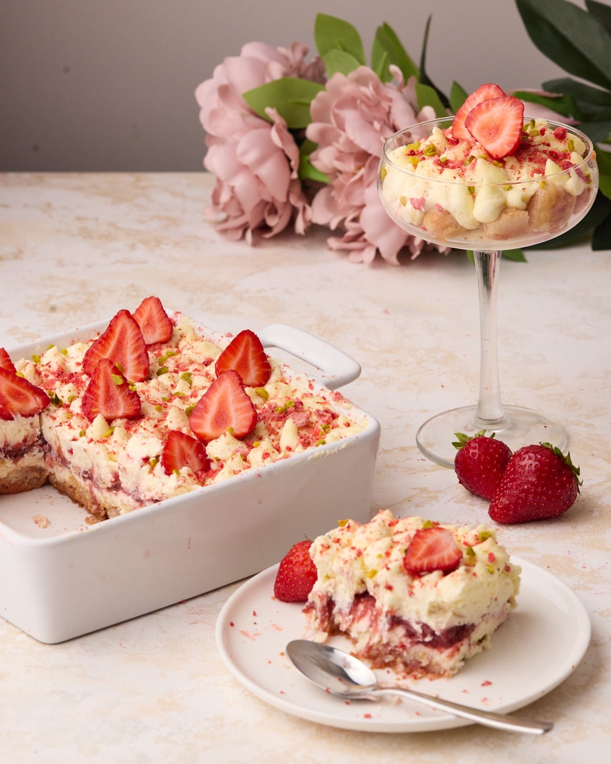 Table with a dish of strawberry tiramisu with one slice next to it on a plate, and another individual portion in a coupe glass behind it.