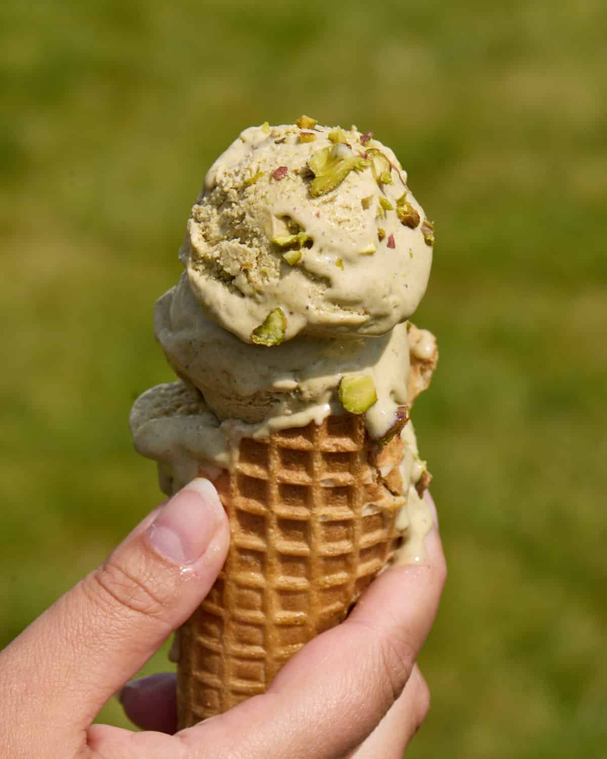 Three scoops of homemade pistachios ice cream piled high in a waffle cone with chopped pistachios sprinkled on top.